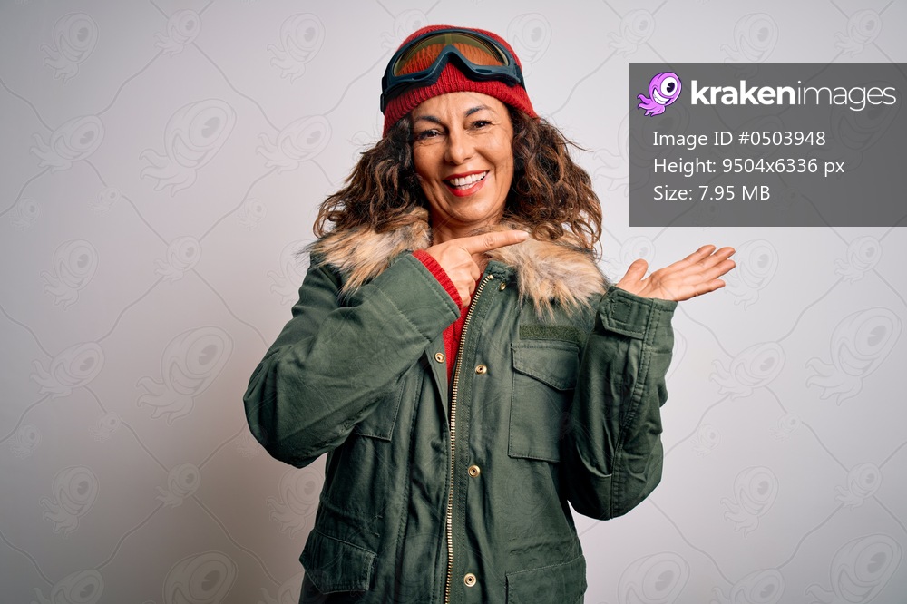 Middle age skier woman wearing snow sportswear and ski goggles over white background amazed and smiling to the camera while presenting with hand and pointing with finger.