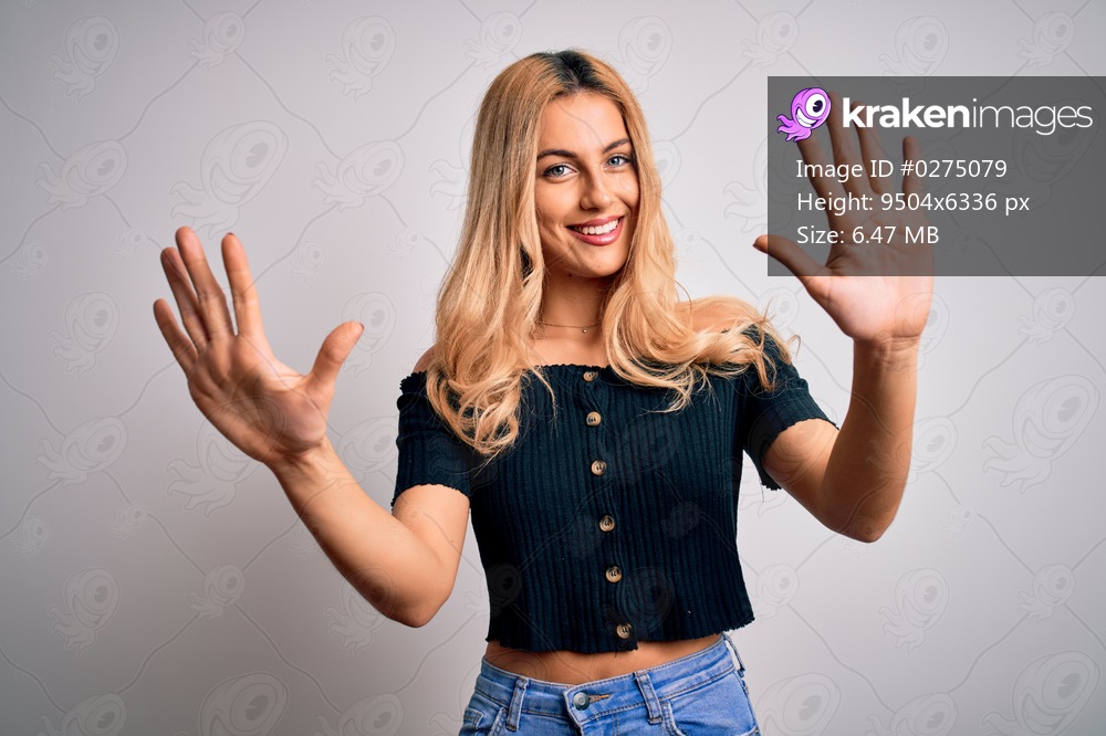 Young beautiful blonde woman wearing casual t-shirt standing over isolated white background showing and pointing up with fingers number ten while smiling confident and happy.