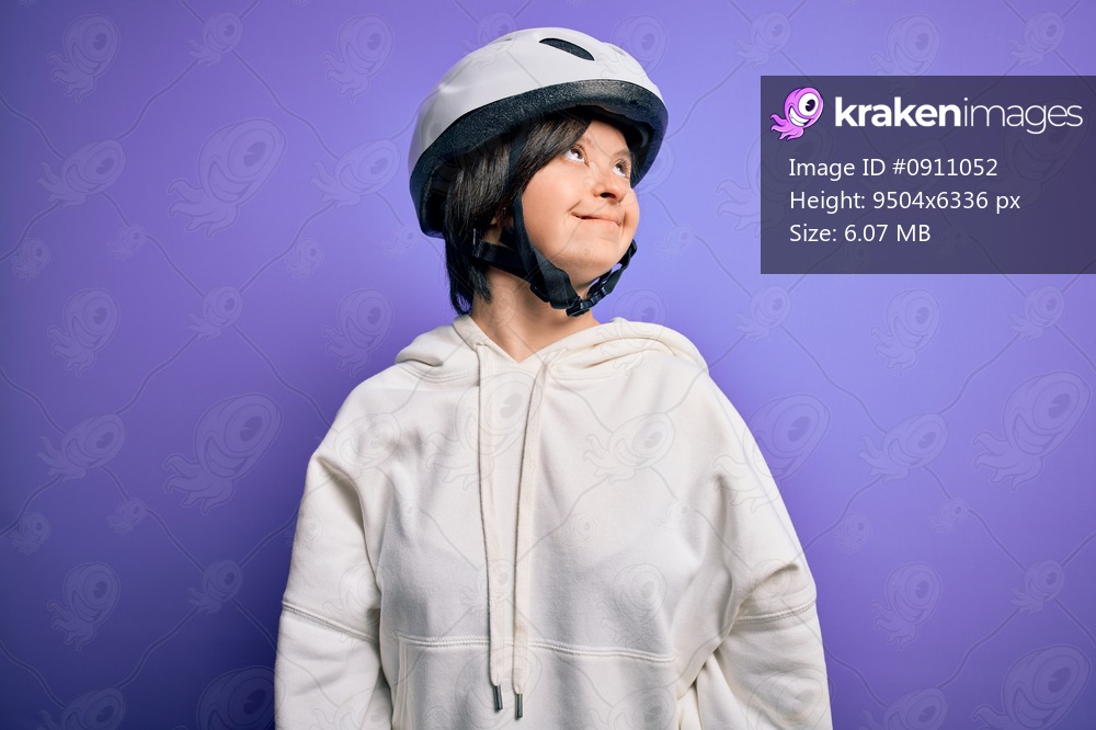 Young down syndrome cyclist woman wearing security bike helmet over purple background looking away to side with smile on face, natural expression. Laughing confident.