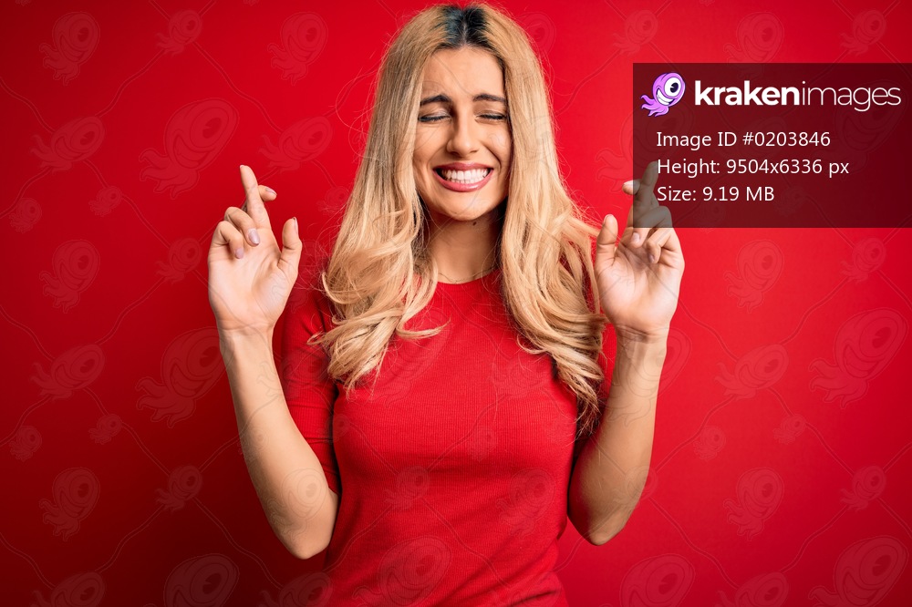 Young beautiful blonde woman wearing casual t-shirt standing over isolated red background gesturing finger crossed smiling with hope and eyes closed. Luck and superstitious concept.