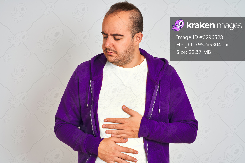 Young man wearing purple sweatshirt standing over isolated white background with hand on stomach because nausea, painful disease feeling unwell. Ache concept.