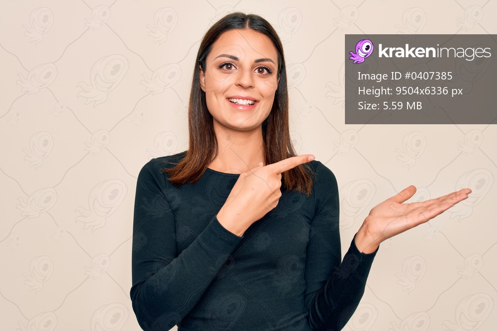 Young beautiful brunette woman wearing casual t-shirt standing over isolated white background amazed and smiling to the camera while presenting with hand and pointing with finger.
