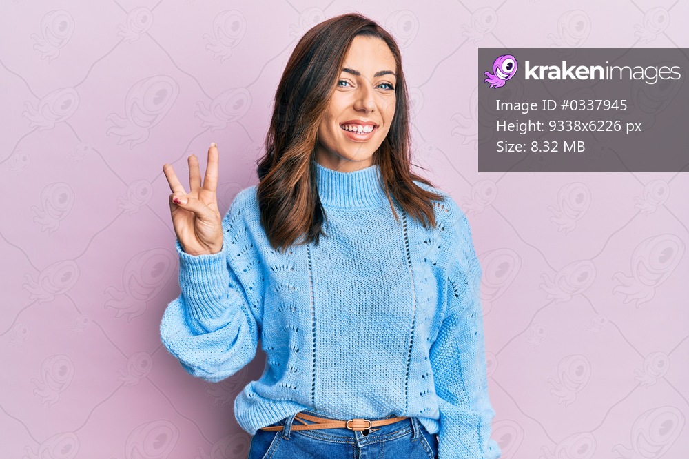 Young brunette woman wearing casual winter sweater over pink background showing and pointing up with fingers number three while smiling confident and happy. 
