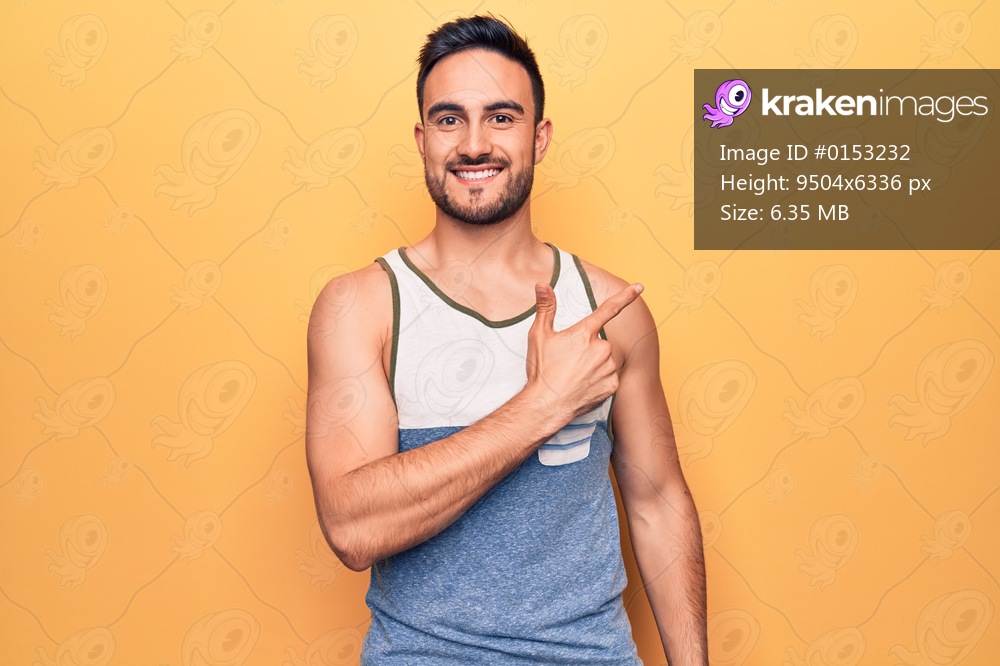 Young handsome man with beard wearing sleeveless t-shirt standing over yellow background smiling cheerful pointing with hand and finger up to the side