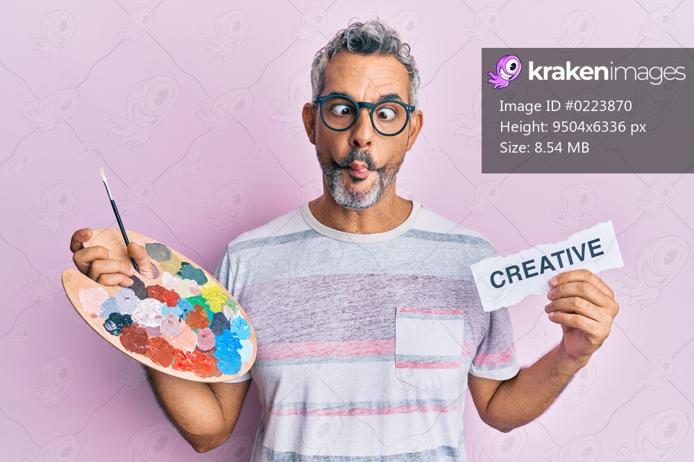 Middle age grey-haired man holding palette and creative message paper making fish face with mouth and squinting eyes, crazy and comical. 