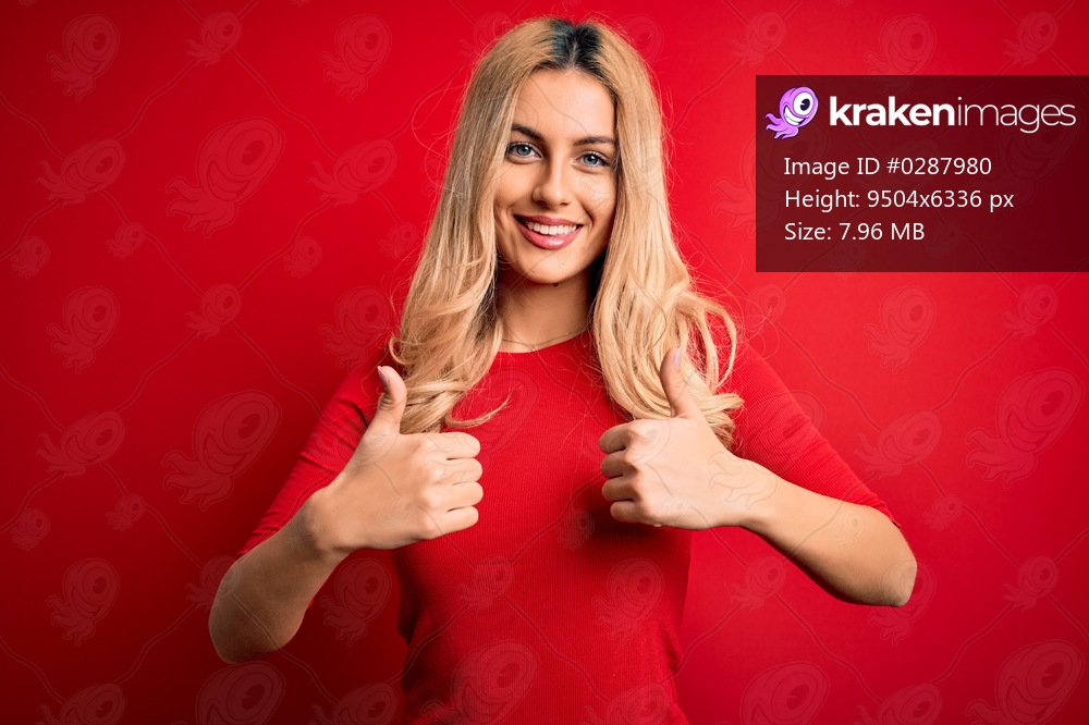 Young beautiful blonde woman wearing casual t-shirt standing over isolated red background success sign doing positive gesture with hand, thumbs up smiling and happy. Cheerful expression and winner gesture.