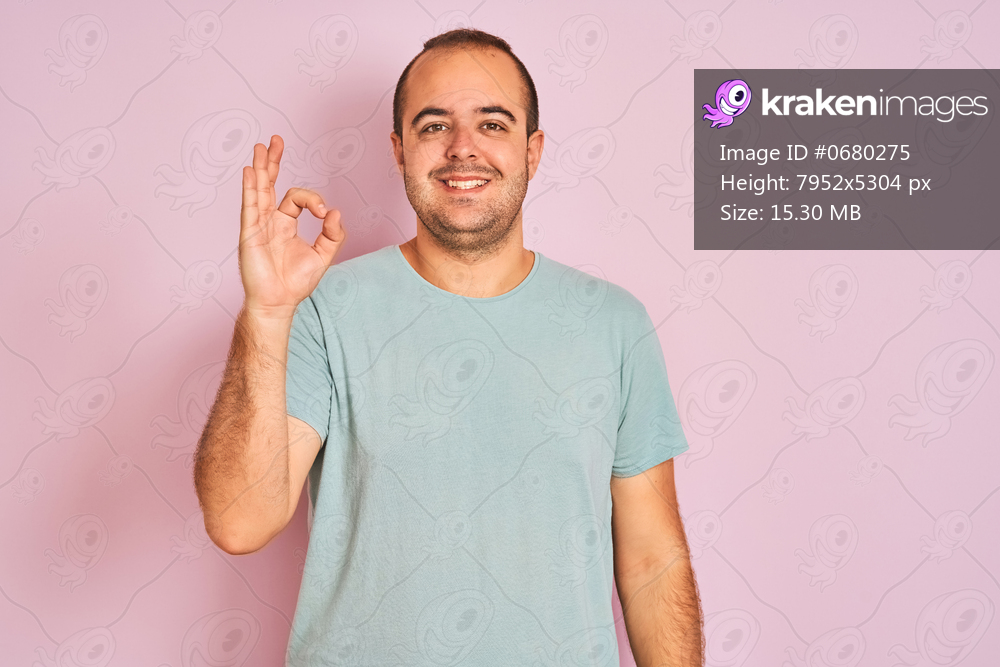 Young man wearing blue casual t-shirt standing over isolated pink background smiling positive doing ok sign with hand and fingers. Successful expression.