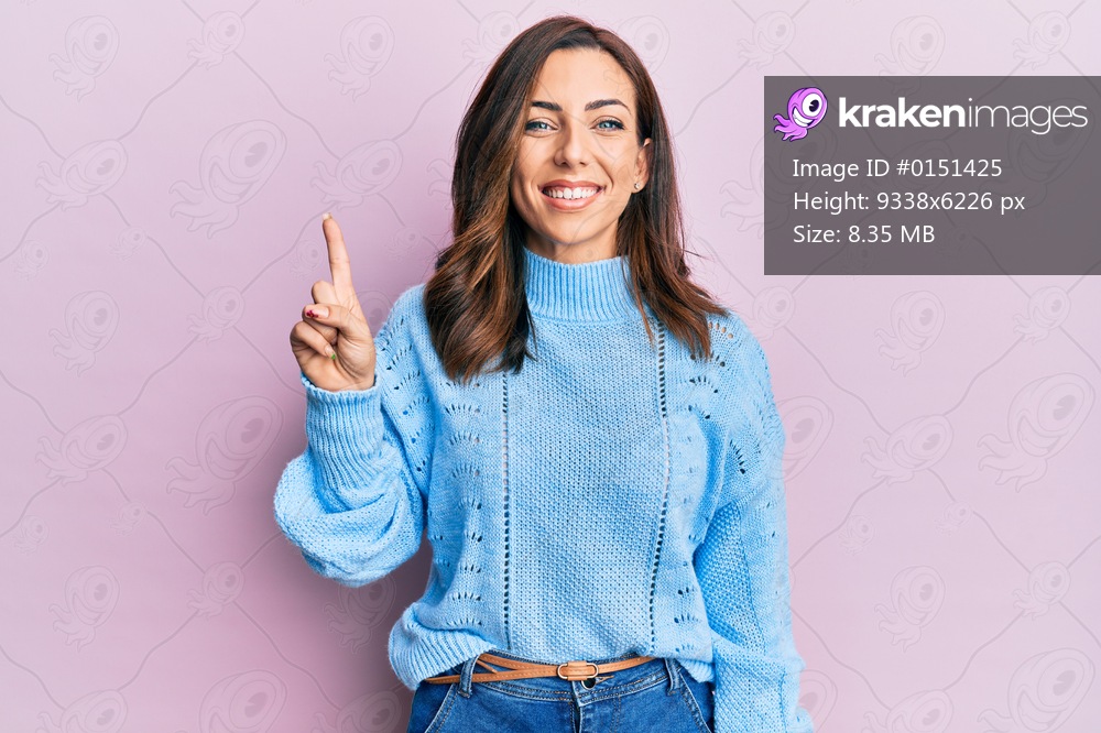 Young brunette woman wearing casual winter sweater over pink background showing and pointing up with finger number one while smiling confident and happy. 