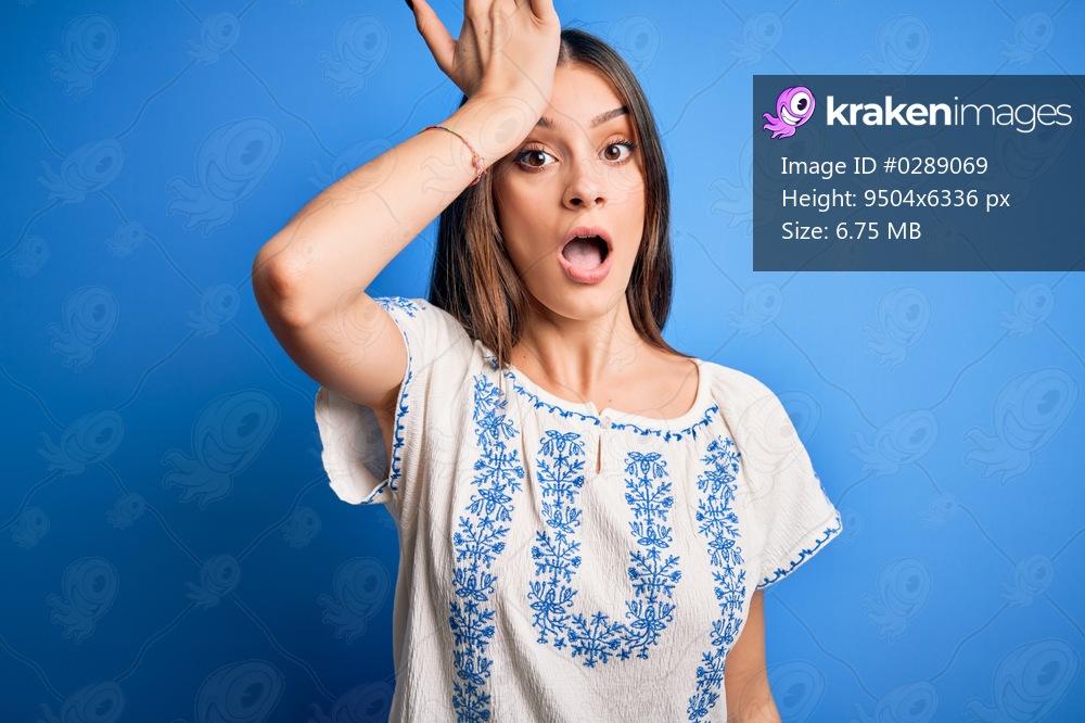 Young beautiful brunette woman wearing casual t-shirt standing over blue background surprised with hand on head for mistake, remember error. Forgot, bad memory concept.