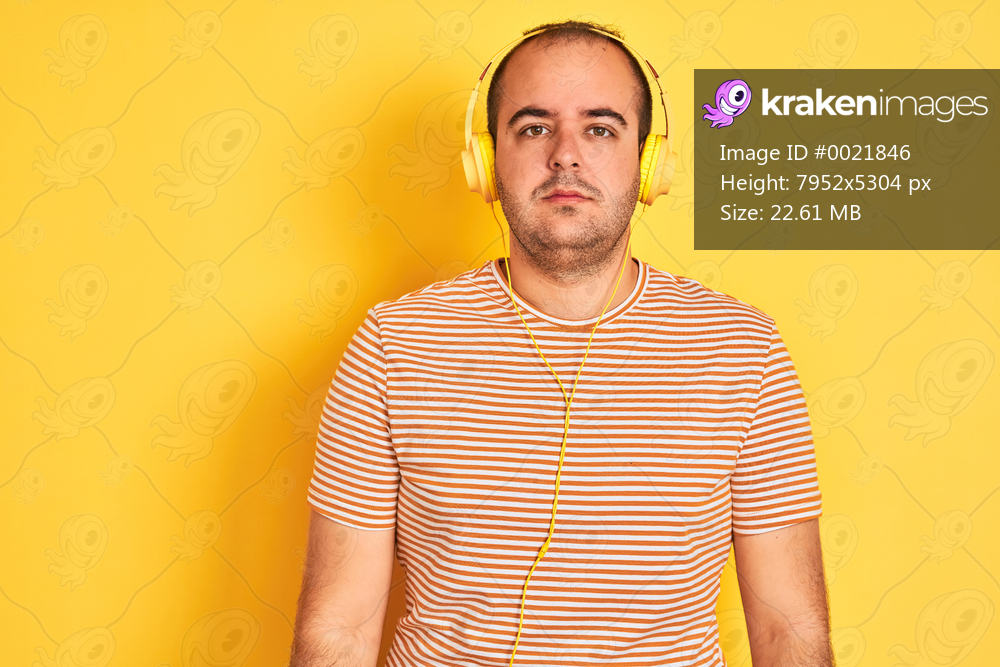 Young man listening to music using headphones standing over isolated yellow background with serious expression on face. Simple and natural looking at the camera.