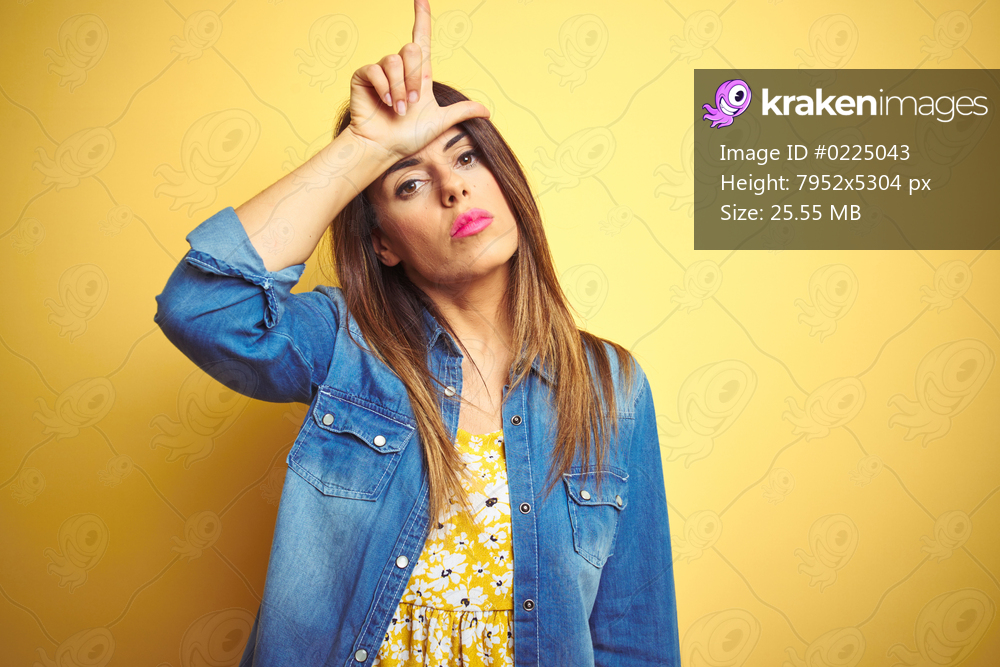 Young beautiful woman standing over yellow isolated background making fun of people with fingers on forehead doing loser gesture mocking and insulting.