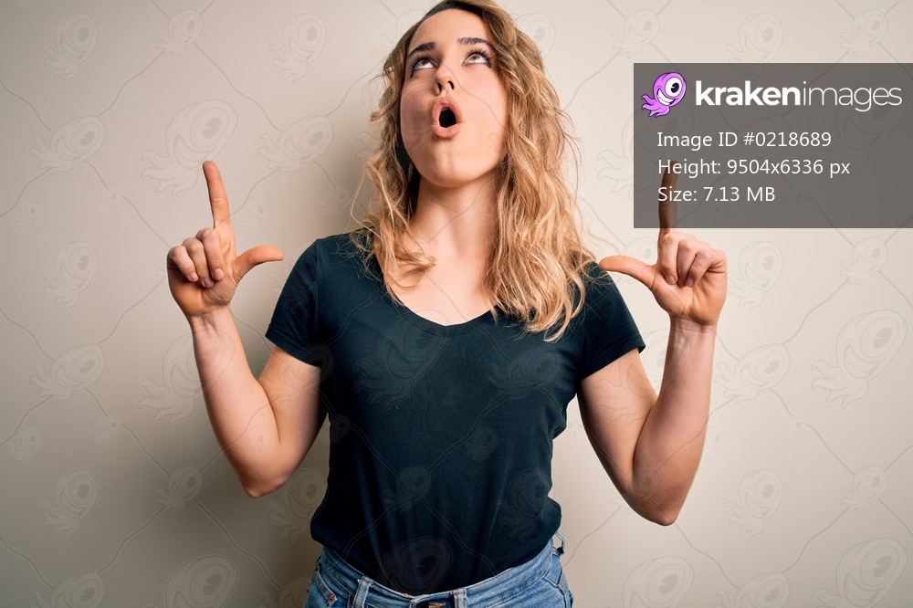Young beautiful blonde woman wearing casual t-shirt standing over isolated white background amazed and surprised looking up and pointing with fingers and raised arms.