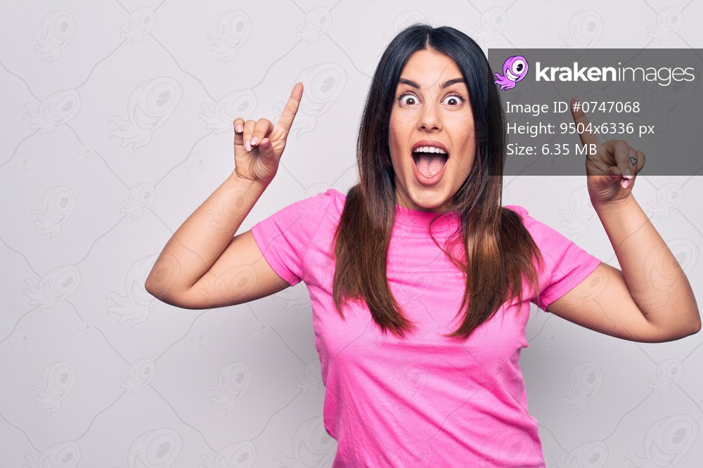 Young beautiful brunette woman wearing casual pink t-shirt standing over white background smiling amazed and surprised and pointing up with fingers and raised arms.
