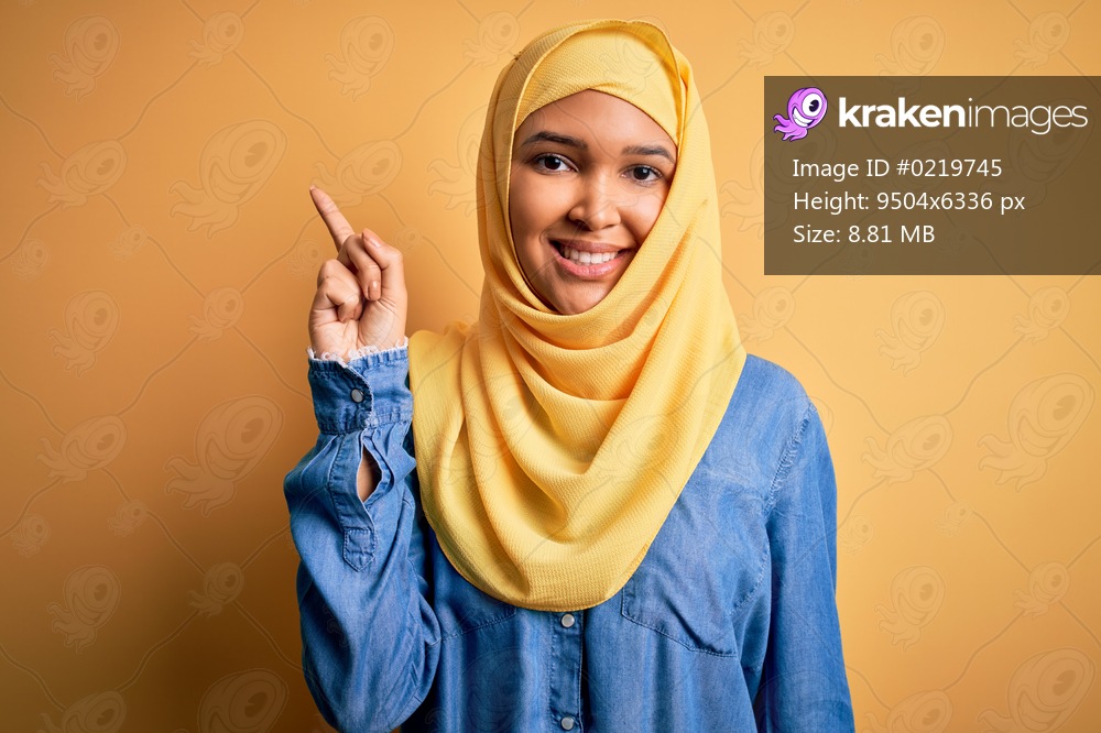 Young beautiful woman with curly hair wearing arab traditional hijab over yellow background pointing finger up with successful idea. Exited and happy. Number one.