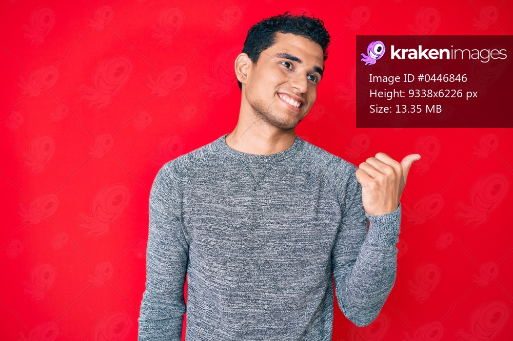 Young handsome hispanic man over red background smiling with happy face looking and pointing to the side with thumb up. 