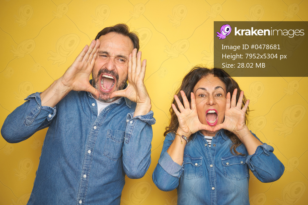 Beautiful middle age couple together standing over isolated yellow background Smiling cheerful playing peek a boo with hands showing face. Surprised and exited