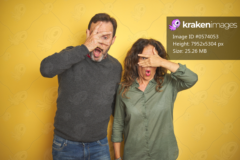 Beautiful middle age couple over isolated yellow background peeking in shock covering face and eyes with hand, looking through fingers with embarrassed expression.