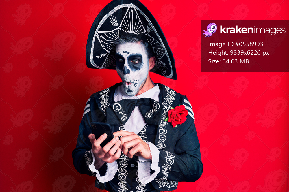 Young man wearing mexican day of the dead costume using smartphone scared and amazed with open mouth for surprise, disbelief face 