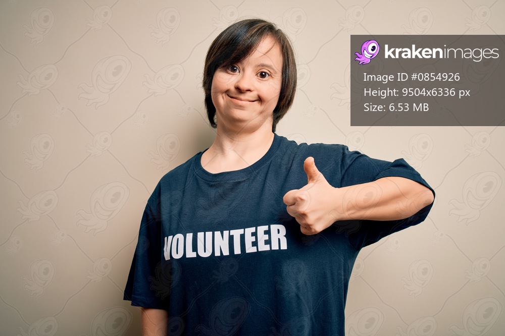 Young down syndrome volunteer woman wearing social care charity t-shirt doing happy thumbs up gesture with hand. Approving expression looking at the camera showing success.
