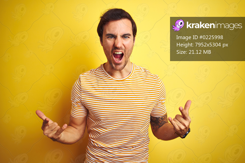 Young handsome man with tattoo wearing striped t-shirt over isolated yellow background shouting with crazy expression doing rock symbol with hands up. Music star. Heavy concept.