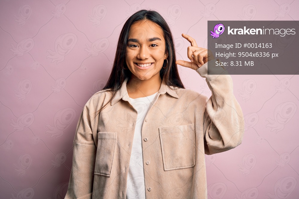 Young beautiful asian woman wearing casual shirt standing over pink background smiling and confident gesturing with hand doing small size sign with fingers looking and the camera. Measure concept.