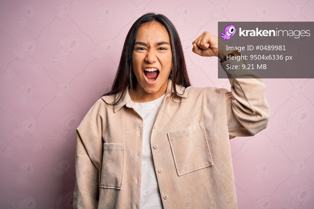 Young beautiful asian woman wearing casual shirt standing over pink background angry and mad raising fist frustrated and furious while shouting with anger. Rage and aggressive concept.