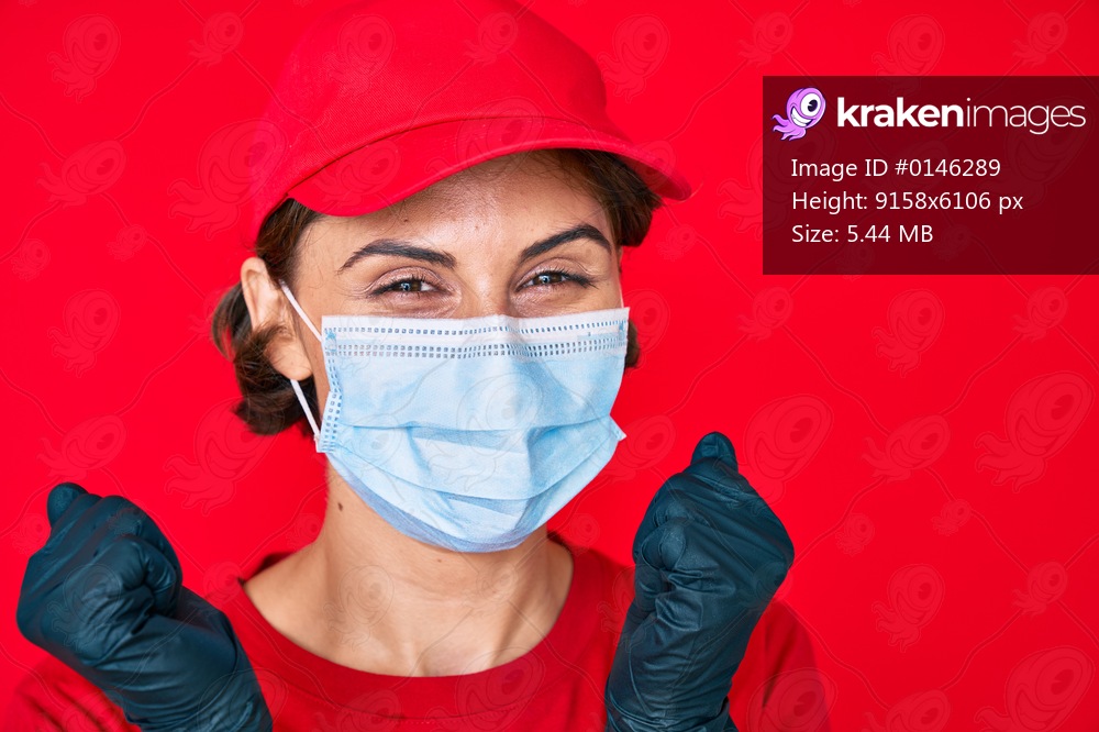 Young hispanic girl wearing delivery uniform and covid-19 safety mask screaming proud, celebrating victory and success very excited with raised arm 