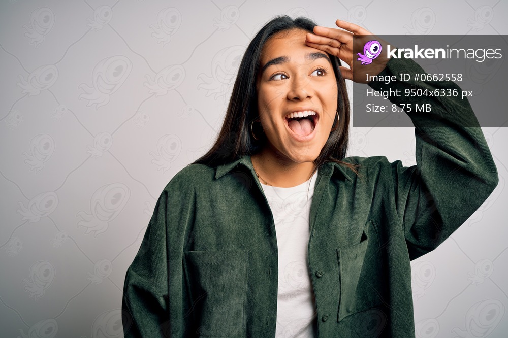 Young beautiful asian woman wearing casual shirt standing over isolated white background very happy and smiling looking far away with hand over head. Searching concept.