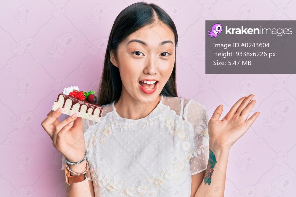 Young chinese woman eating a tasty cake celebrating achievement with happy smile and winner expression with raised hand 