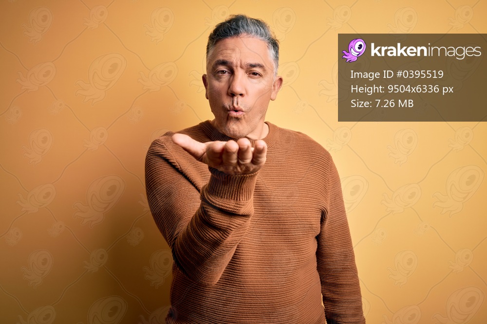 Middle age handsome grey-haired man wearing casual sweater over yellow background looking at the camera blowing a kiss with hand on air being lovely and sexy. Love expression.