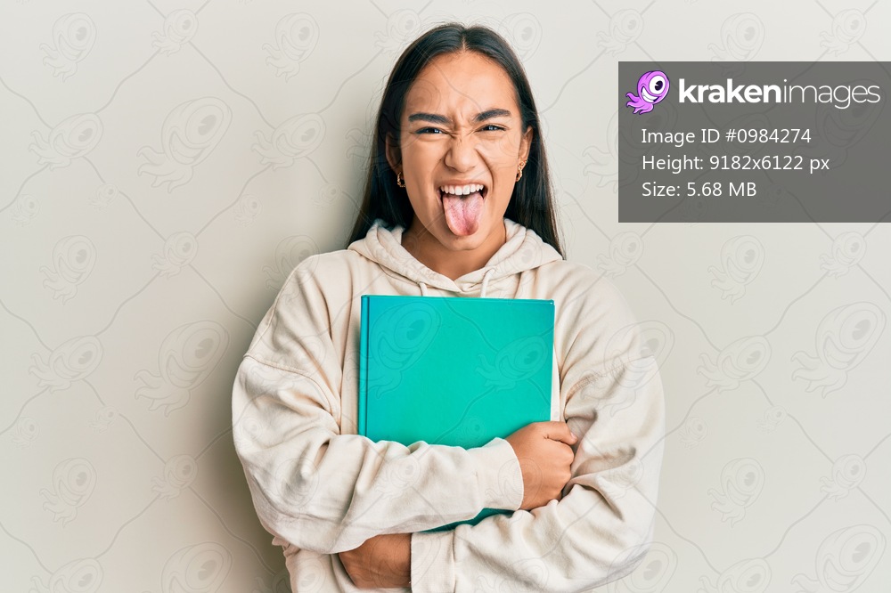 Young asian woman holding book sticking tongue out happy with funny expression. 