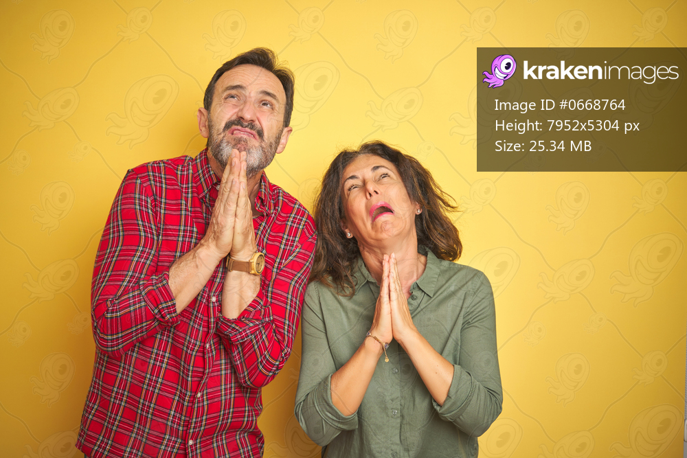 Beautiful middle age couple over isolated yellow background begging and praying with hands together with hope expression on face very emotional and worried. Asking for forgiveness. Religion concept.