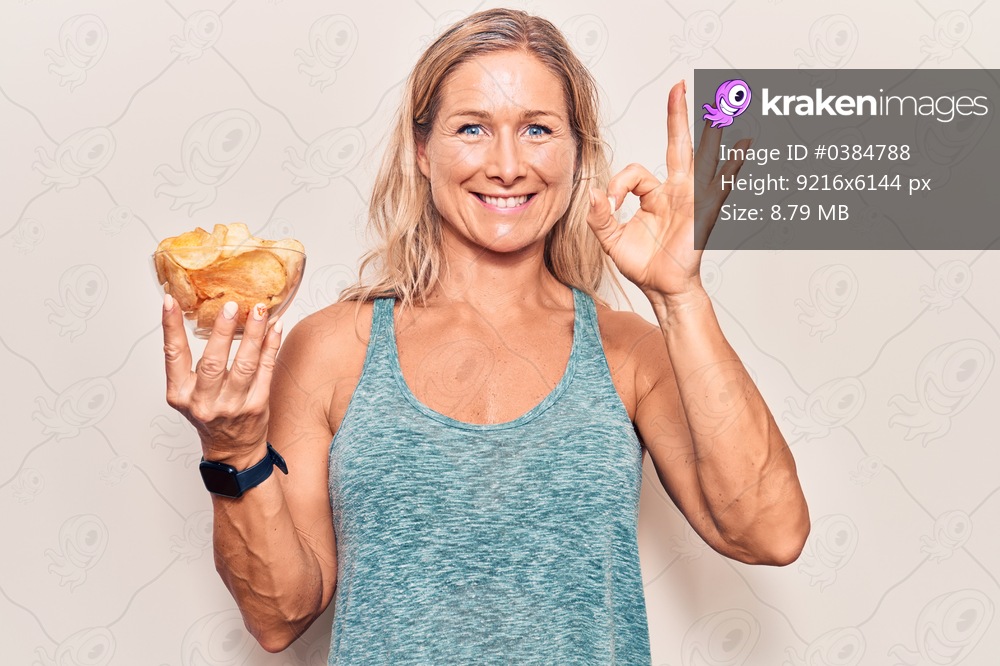 Middle age caucasian blonde woman holding potato chips doing ok sign with fingers, smiling friendly gesturing excellent symbol 