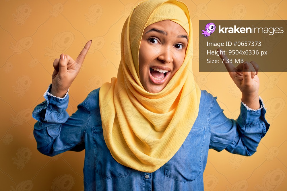 Young beautiful woman with curly hair wearing arab traditional hijab over yellow background smiling amazed and surprised and pointing up with fingers and raised arms.