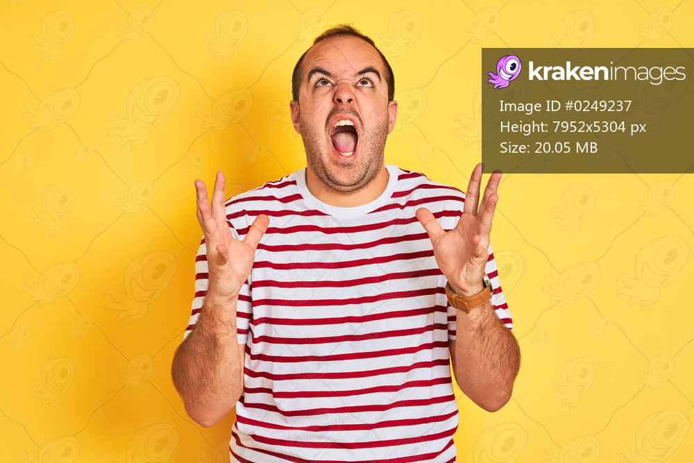 Young man wearing casual striped t-shirt standing over isolated yellow background crazy and mad shouting and yelling with aggressive expression and arms raised. Frustration concept.