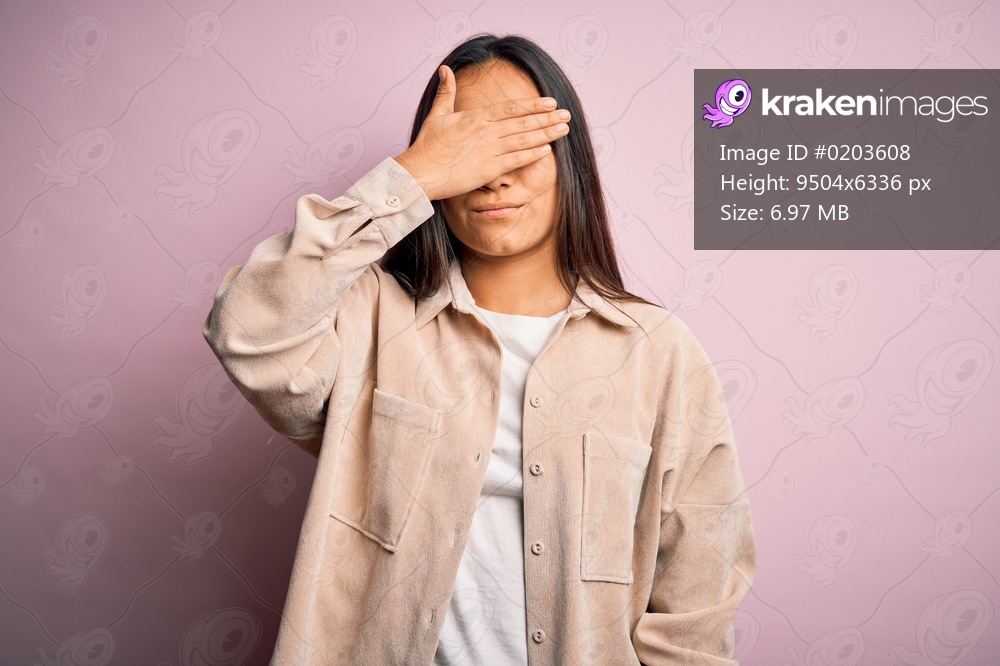 Young beautiful asian woman wearing casual shirt standing over pink background covering eyes with hand, looking serious and sad. Sightless, hiding and rejection concept