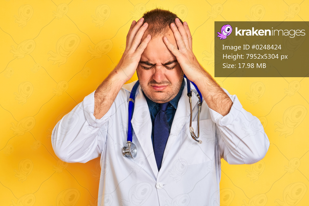 Young doctor man wearing coat and stethoscope standing over isolated yellow background suffering from headache desperate and stressed because pain and migraine. Hands on head.