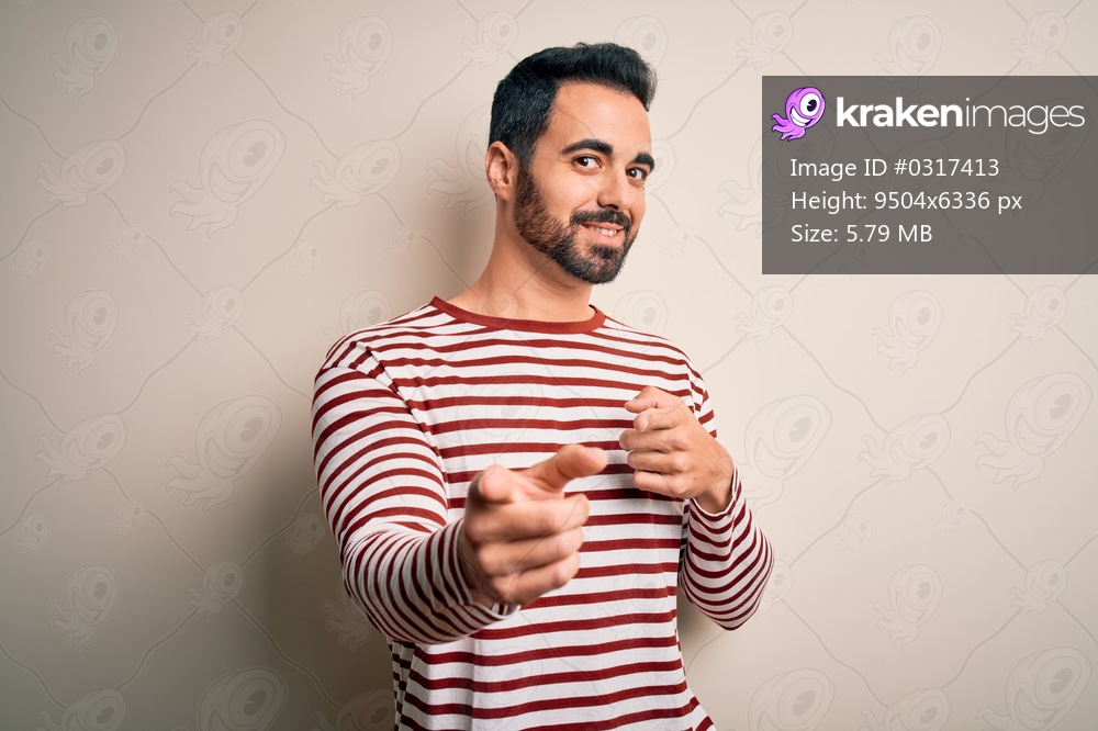 Young handsome man with beard wearing casual striped t-shirt standing over white background pointing fingers to camera with happy and funny face. Good energy and vibes.