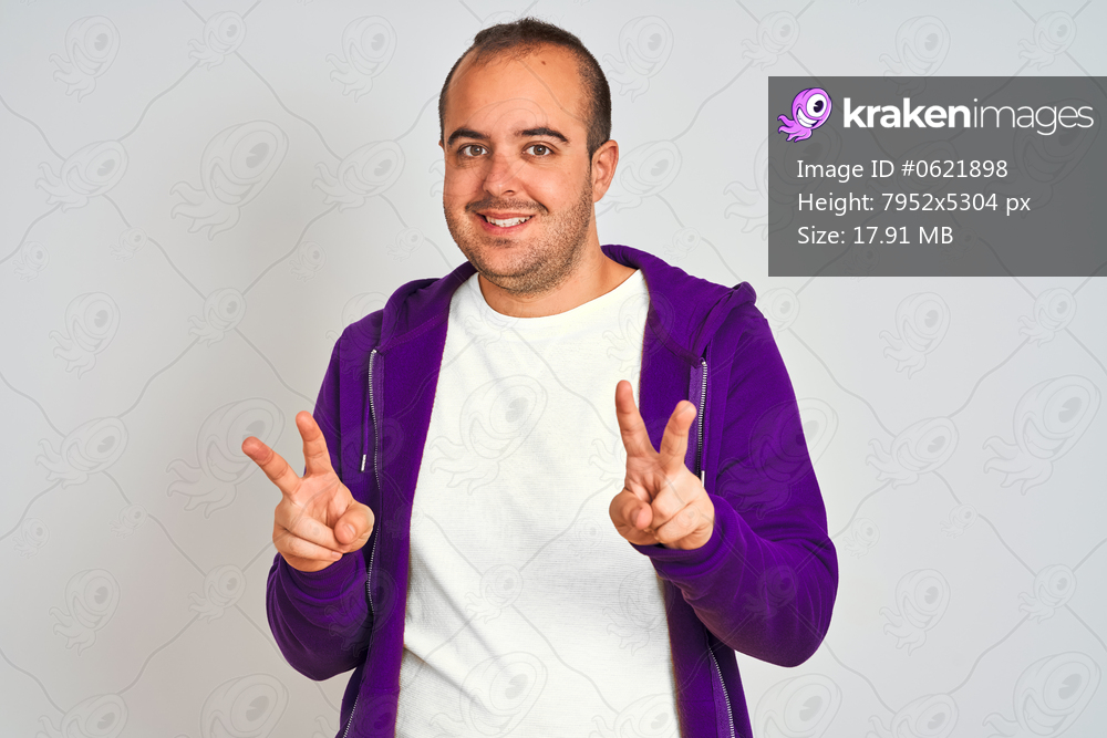 Young man wearing purple sweatshirt standing over isolated white background smiling looking to the camera showing fingers doing victory sign. Number two.