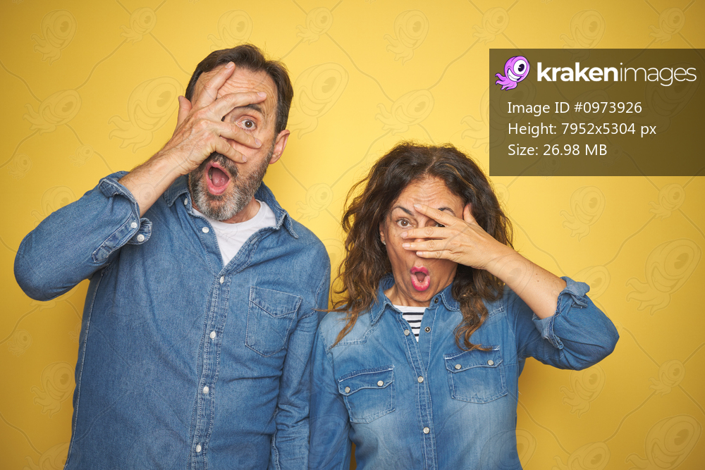 Beautiful middle age couple together standing over isolated yellow background peeking in shock covering face and eyes with hand, looking through fingers with embarrassed expression.