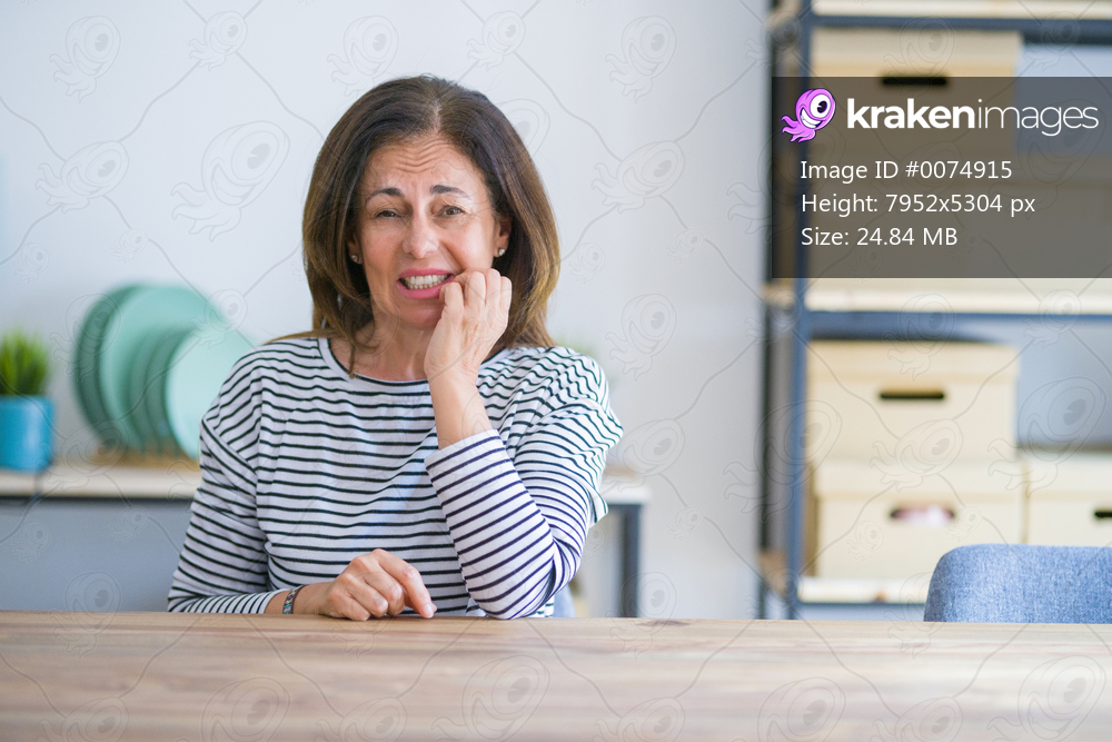 Middle age senior woman sitting at the table at home looking stressed and nervous with hands on mouth biting nails. Anxiety problem.