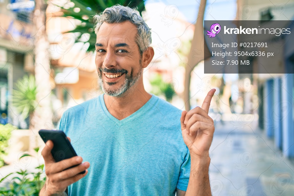 Middle age grey-haired man using smartphone at street of city smiling happy pointing with hand and finger to the side 