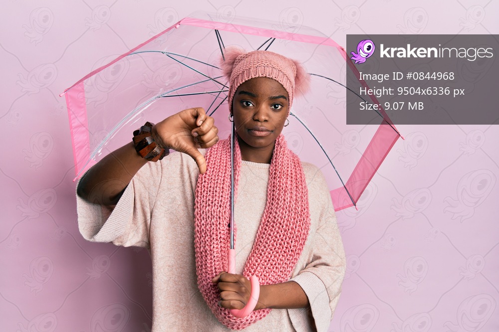 Young african american woman holding umbrella with angry face, negative sign showing dislike with thumbs down, rejection concept 