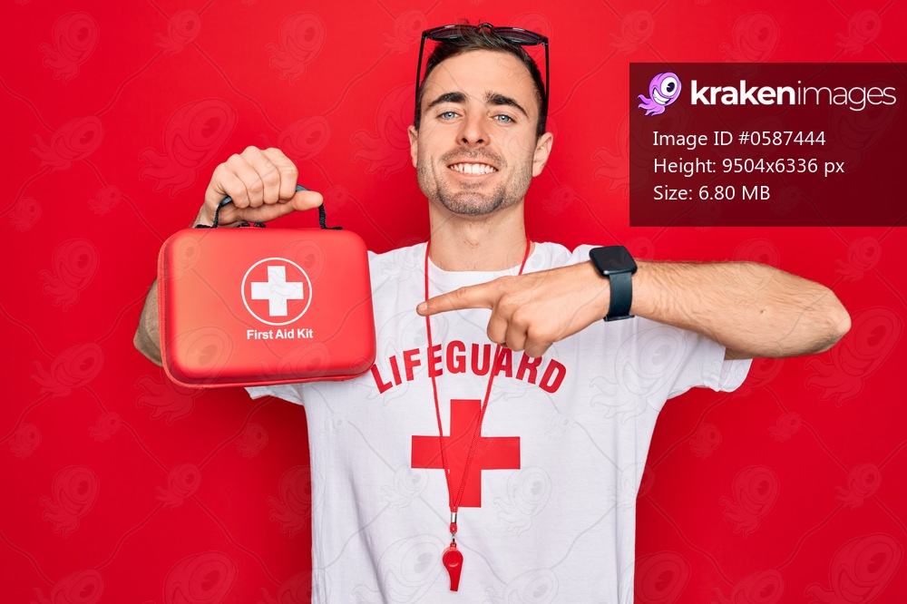 Young handsome lifeguard man wearing t-shirt with red cross and whistle holding first aid kit very happy pointing with hand and finger