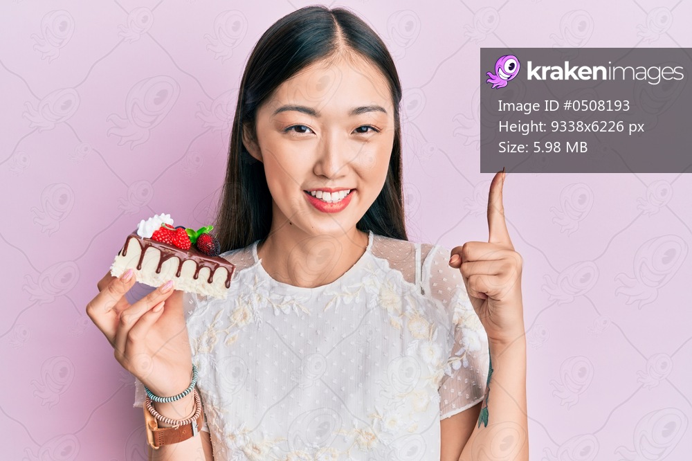 Young chinese woman eating a tasty cake smiling with an idea or question pointing finger with happy face, number one 