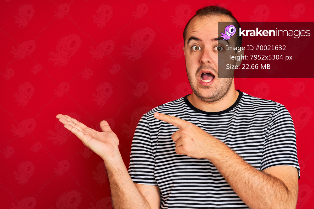 Young man wearing striped navy t-shirt standing over isolated red background amazed and smiling to the camera while presenting with hand and pointing with finger.