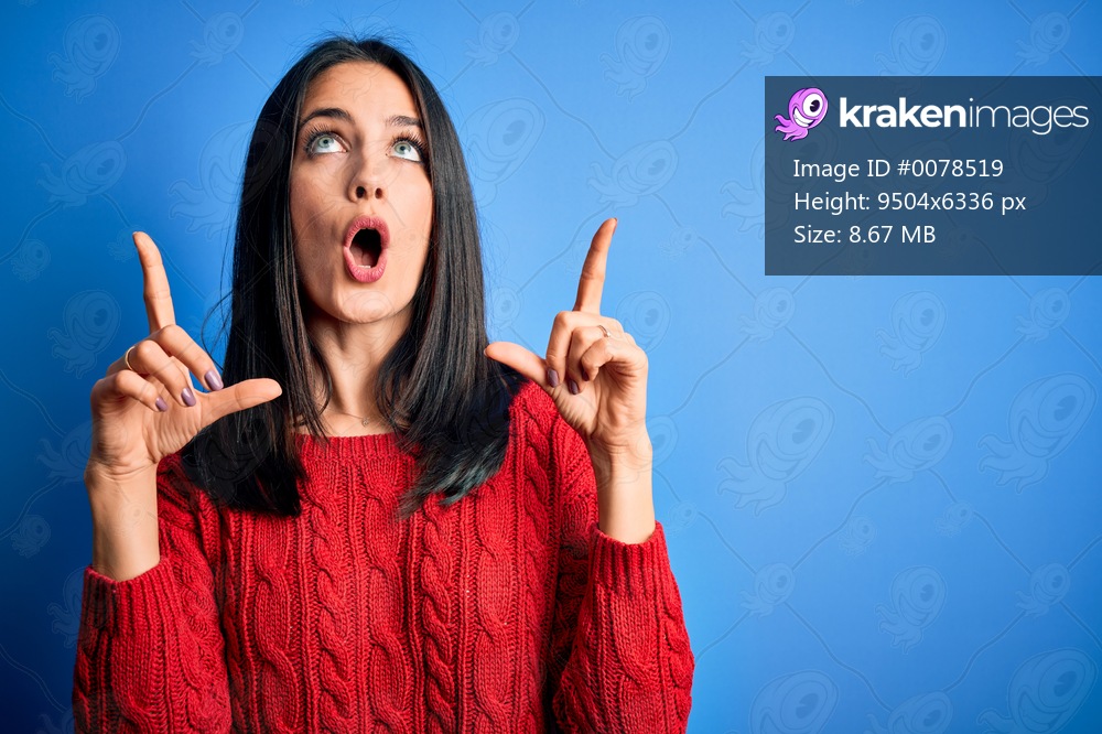 Young brunette woman with blue eyes wearing red casual sweater over isolated background amazed and surprised looking up and pointing with fingers and raised arms.