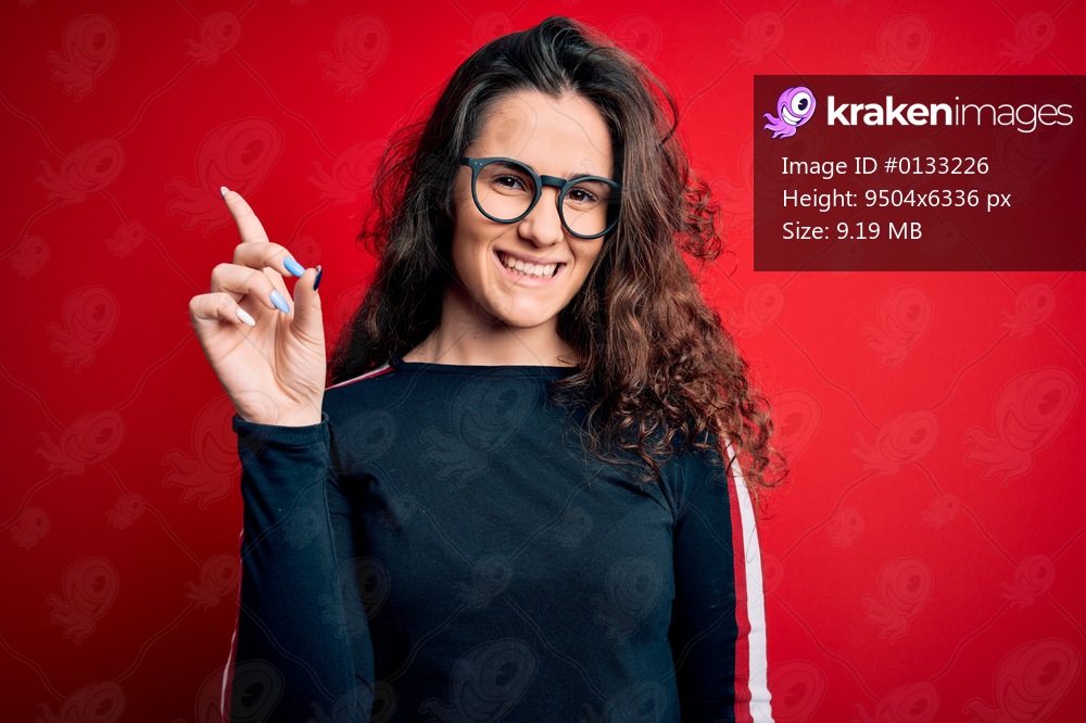 Young beautiful woman with curly hair wearing sweater and glasses over red background showing and pointing up with finger number one while smiling confident and happy.