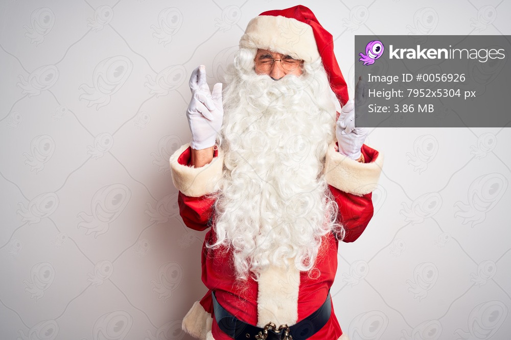 Middle age handsome man wearing Santa costume standing over isolated white background gesturing finger crossed smiling with hope and eyes closed. Luck and superstitious concept.
