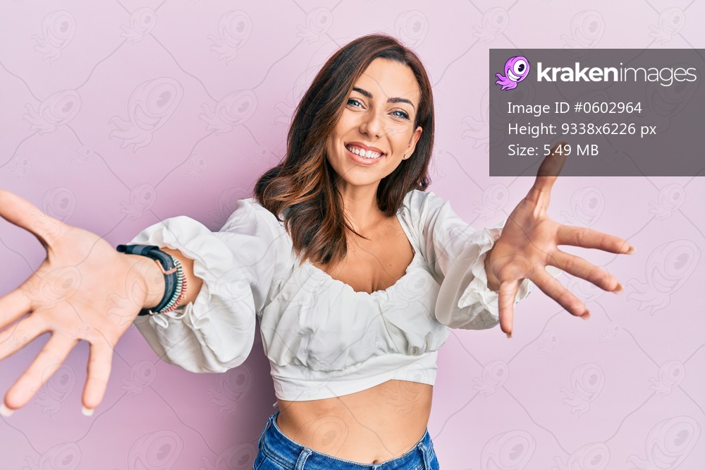 Young brunette woman wearing casual clothes over pink looking at the camera smiling with open arms for hug. cheerful expression embracing happiness. 